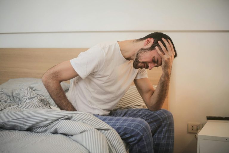 Image of a man sitting on the edge of a bed with his head in his left hand looking in pain